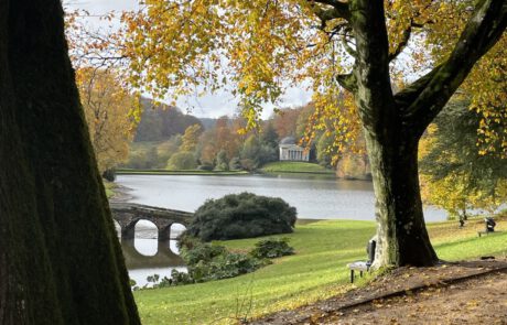 Das Bild zeigt einen Park im Herbst. Im Vordergrund befindet sich ein Baumstamm. Ein Kiesweg ist mit gelbem Laub bedeckt. Über einen See führt eine Brücke aus Stein. Am gegenüberliegenden Seeufer befindet sich ein weißes Gebäude. Die Bäume im Hintergrund haben herbstlich gefärbtes Laub.