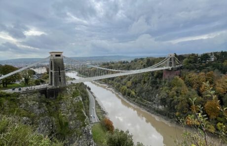 Eine Hängebrücke erstreckt sich von einer Seite einer Schlucht zur anderen. unter ihr fließt ein Fluss. Das Wasser ist braun. An den Hängen der Schlucht wachten Bäume mit buntem Herbstlaub. Der Himmel ist bewölkt.