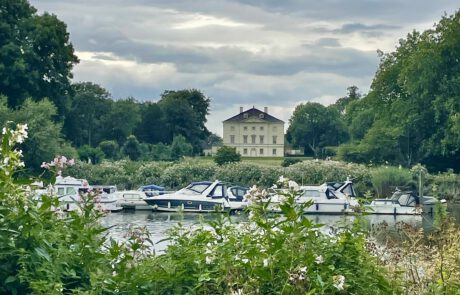 Blick über die Themse bei London Richtung Marble Hill House in Richmond. Das Haus ist hell farbig, eingebettet in einen Park mit großen Bäumen. Im Vordergrund befinden sich Motorboote auf der Themse.